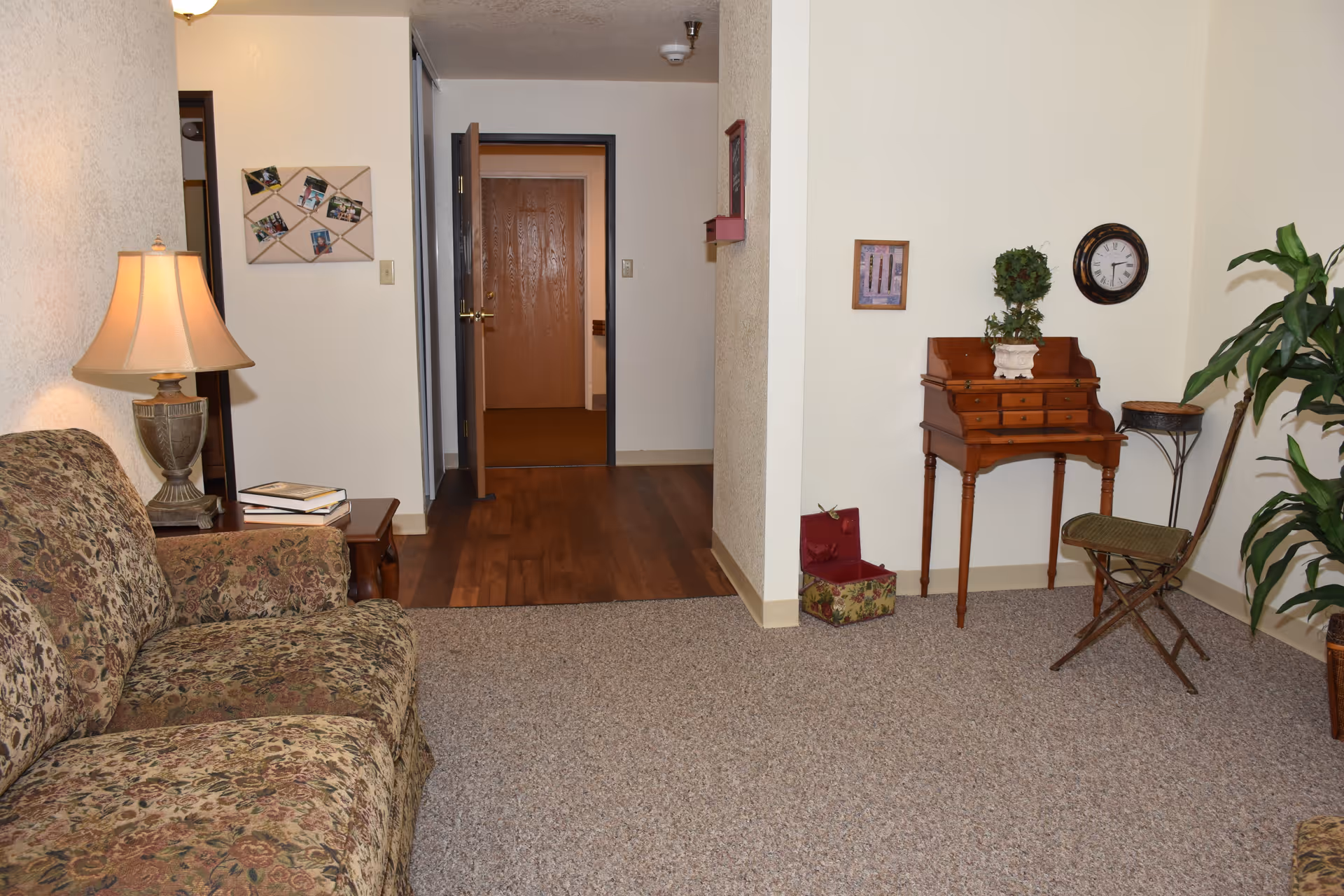 Cozy living room with a patterned sofa, side table and lamp, a small writing desk and plants, and an open doorway to a hallway.