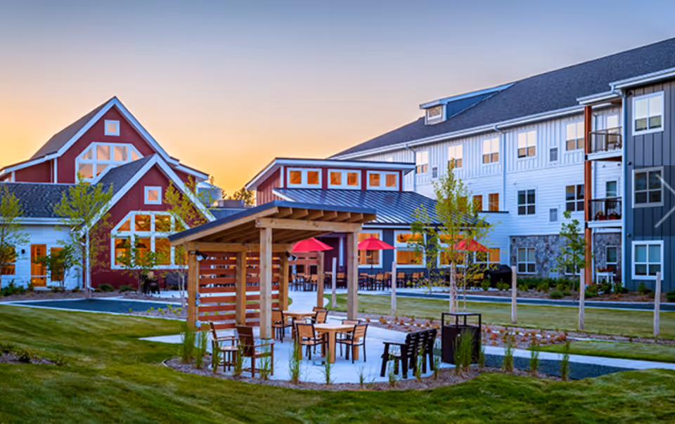 Outdoor seating area with wooden pergola and tables surrounded by chairs on a concrete patio, with a large multi-story building and a barn-style building in the background during sunset.