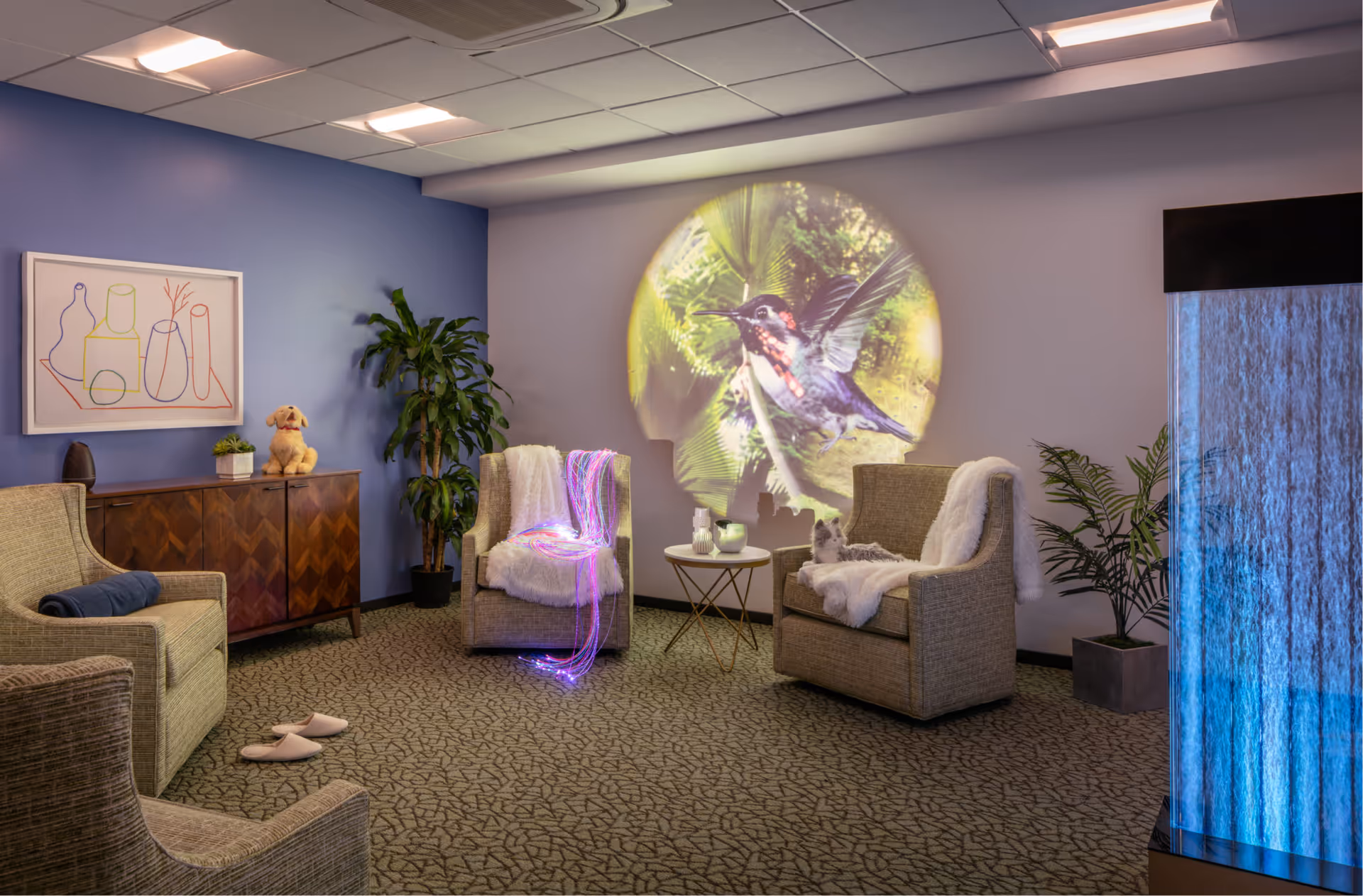 A cozy sitting area with four cushioned armchairs arranged in a semi-circle on a patterned carpet. Two chairs have white fluffy throws, one with colorful fiber optic strands and the other with a small stuffed animal. A small round table with decorative items is between the two chairs. On the wall, a large circular projection of a hummingbird with green foliage is displayed. A wooden cabinet with a small plant and a stuffed dog is against a blue wall, and there are two potted plants in the room. A tall water feature with blue lighting is on the right side.