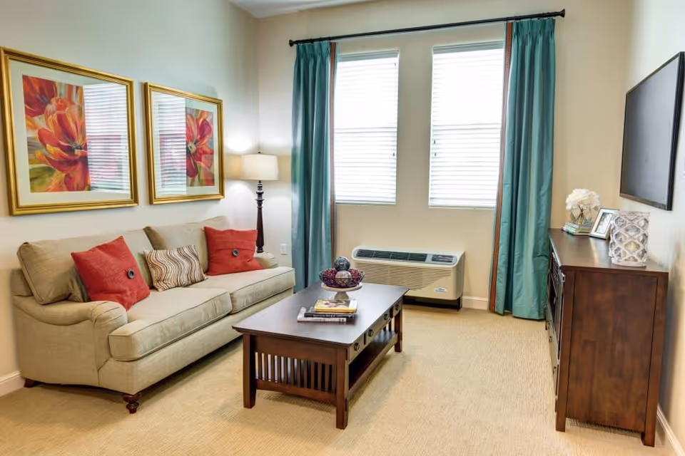 A cozy living room with a beige sofa adorned with red and patterned cushions, a wooden coffee table with decorative items and books, two framed floral paintings on the wall, a floor lamp, teal curtains framing two windows with blinds, and a wooden cabinet with decorative items and a flat-screen TV mounted above it.