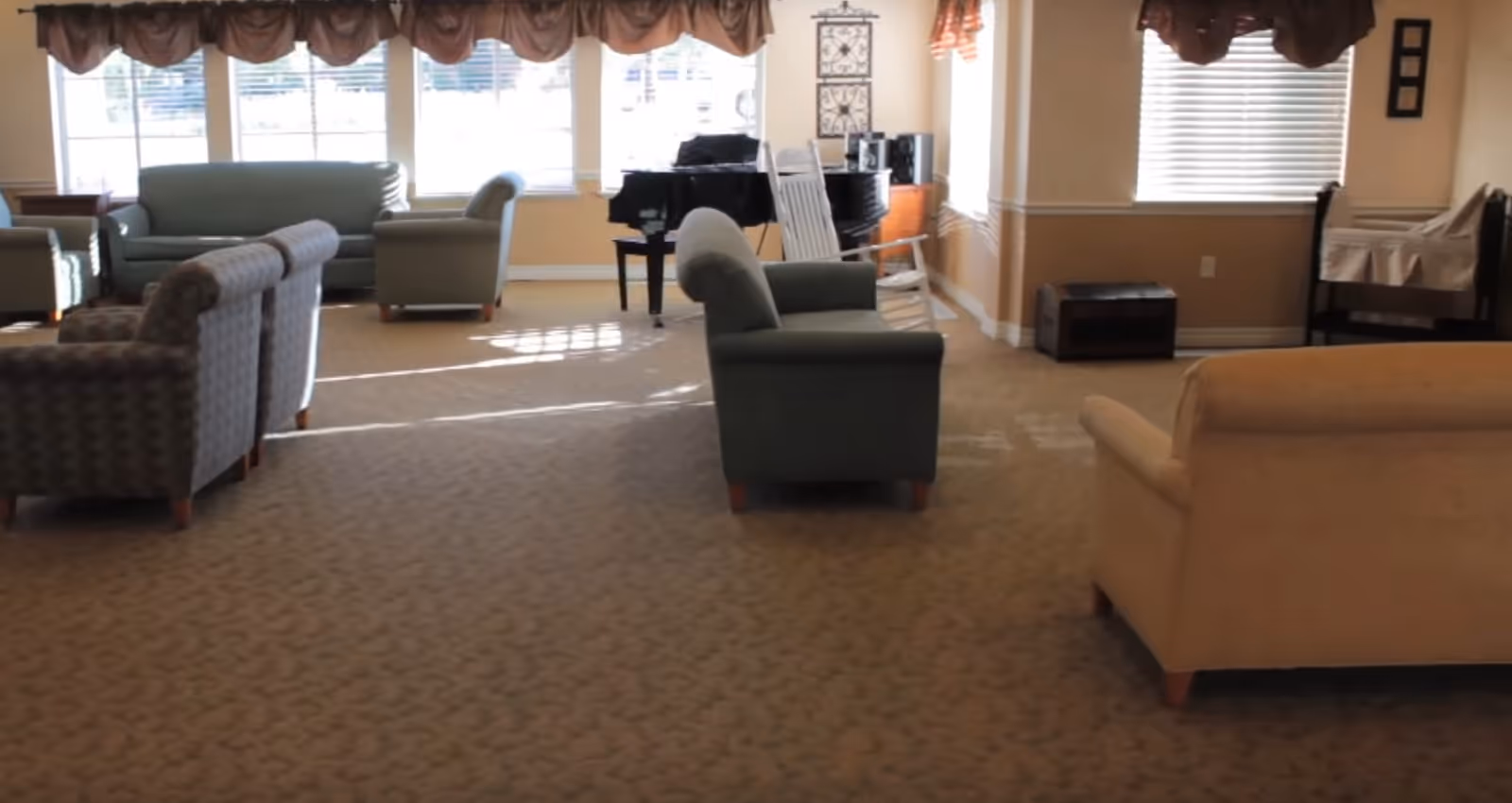 A spacious living room area with multiple armchairs and sofas arranged on a patterned carpet. Large windows with valances allow natural light to fill the room. A black grand piano and a white rocking chair are positioned near the windows. The walls are painted beige with white trim, and there are decorative wall hangings and a small wooden chest in the corner.