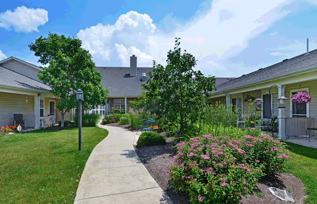 A sunny outdoor courtyard at a senior living facility with a concrete walkway winding through green grass, flowering bushes, and small trees. The courtyard is surrounded by single-story buildings with beige siding and windows. There are benches and hanging flower baskets along the building walls.