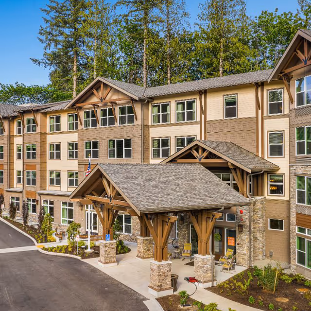 Exterior view of a multi-story senior living facility with a covered entrance supported by wooden beams and stone pillars. The building has numerous windows and is surrounded by landscaped greenery and tall trees in the background.