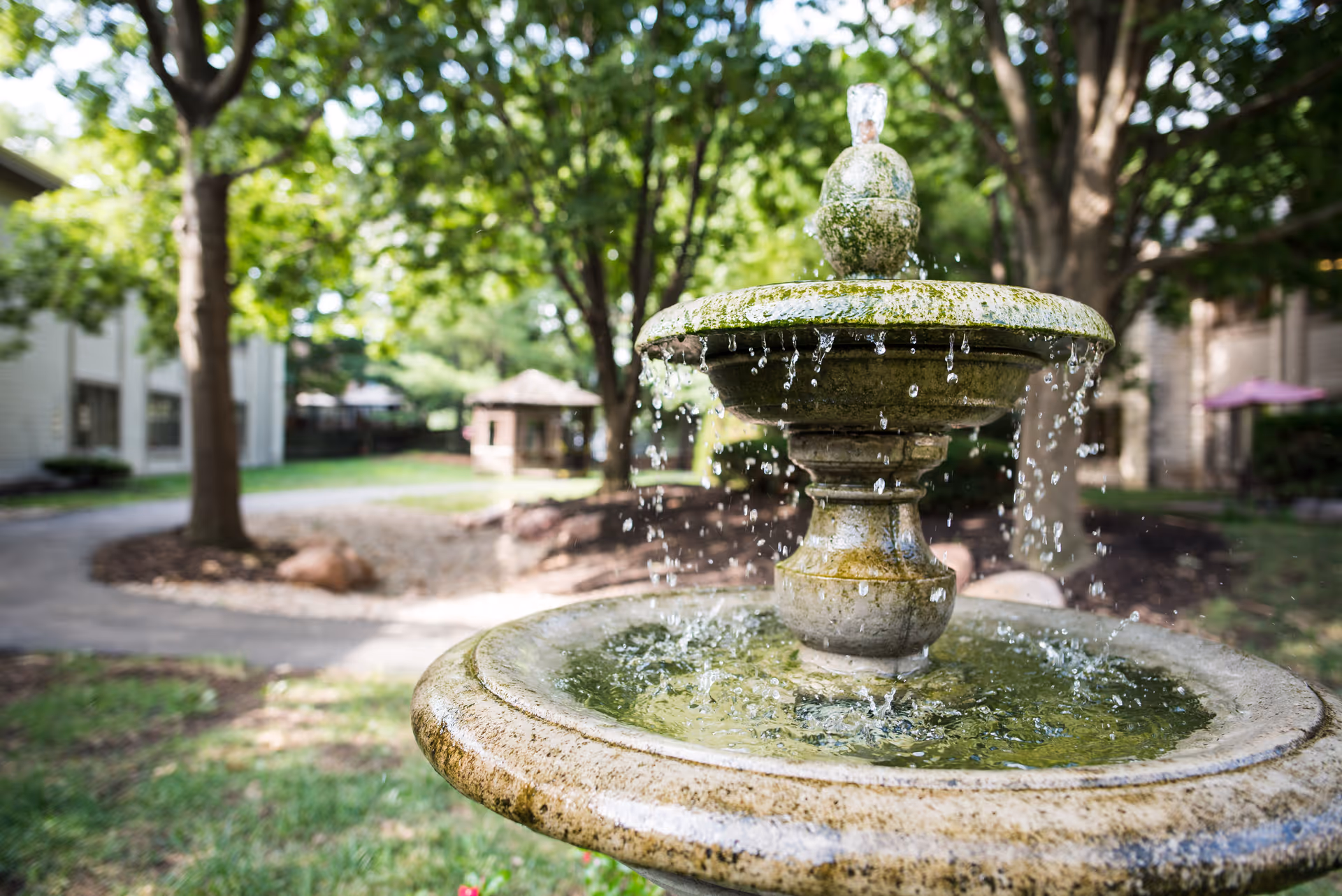 A close-up view of a stone water fountain with water flowing from the top tier to the basin below, set in a green outdoor garden area with trees, grass, and a pathway in the background.