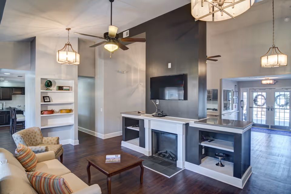 A modern living room area with a beige sofa and two armchairs, a wooden coffee table, a wall-mounted flat-screen TV above a white fireplace with black accents, ceiling fans, and pendant lights. The room has dark wood flooring and light gray walls, with a view into a kitchen area and an entrance with glass doors decorated with wreaths.