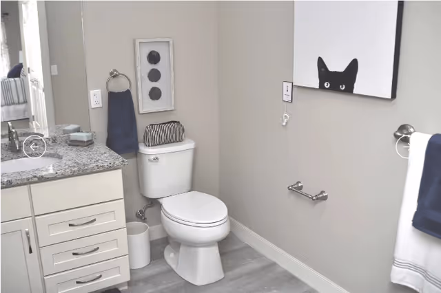 A clean and modern bathroom with a white toilet, a granite countertop with a sink, white cabinetry, and a small trash bin. The walls are painted light gray, decorated with a framed picture of three black circles and a painting of a black cat peeking over a white background. There are navy blue towels hanging on silver towel rings.