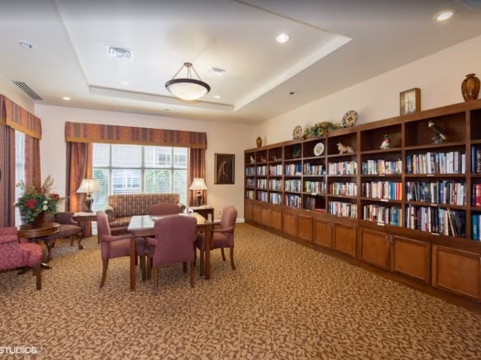 A cozy library or reading room with a large wooden bookshelf filled with books and decorative items along one wall. There is a table with four upholstered chairs in the center of the room, a patterned carpet, and a window with curtains letting in natural light. Two table lamps and a ceiling light fixture provide additional lighting.