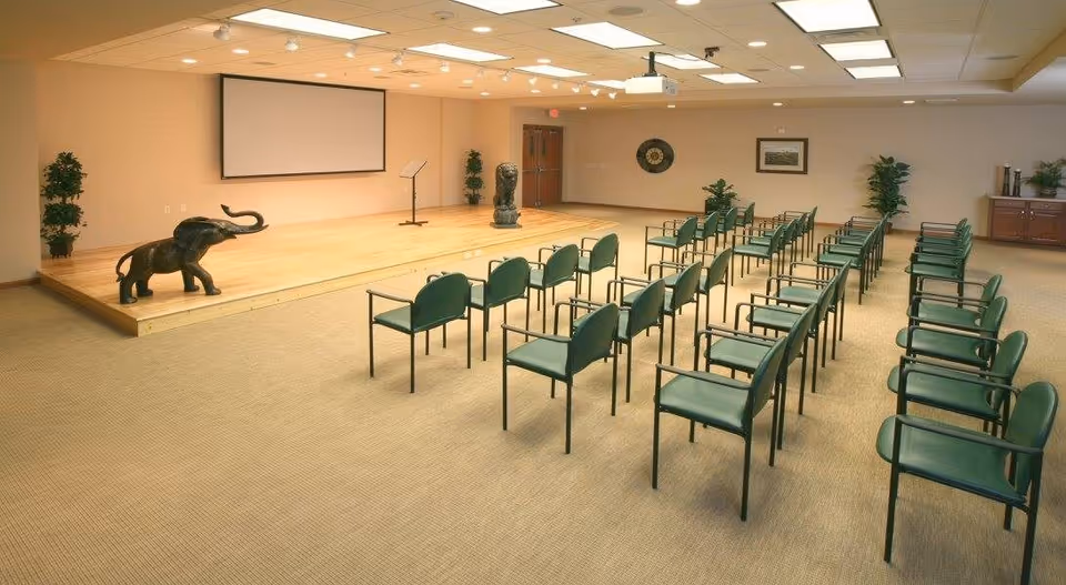A conference or meeting room with rows of green chairs facing a wooden stage. The stage has a music stand, two elephant statues, and a large projection screen on the wall behind it. The room is well-lit with ceiling lights and has beige walls and carpet. There are plants and framed artwork on the walls.