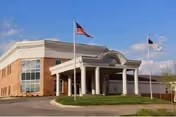 Front entrance of a brick building with a covered portico supported by white columns and flagpoles flying an American flag and another flag against a blue sky.