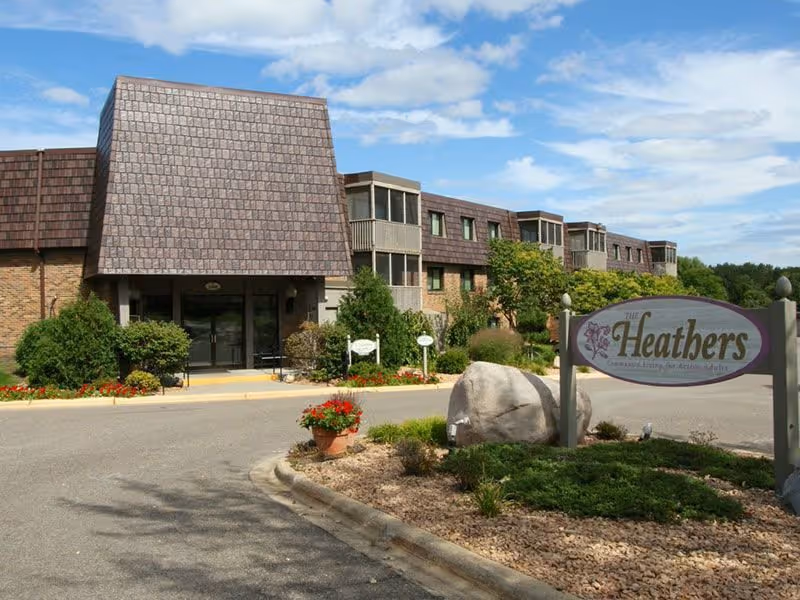 Front entrance of a senior living building with a sign reading "The Heathers", driveway and landscaped planter.