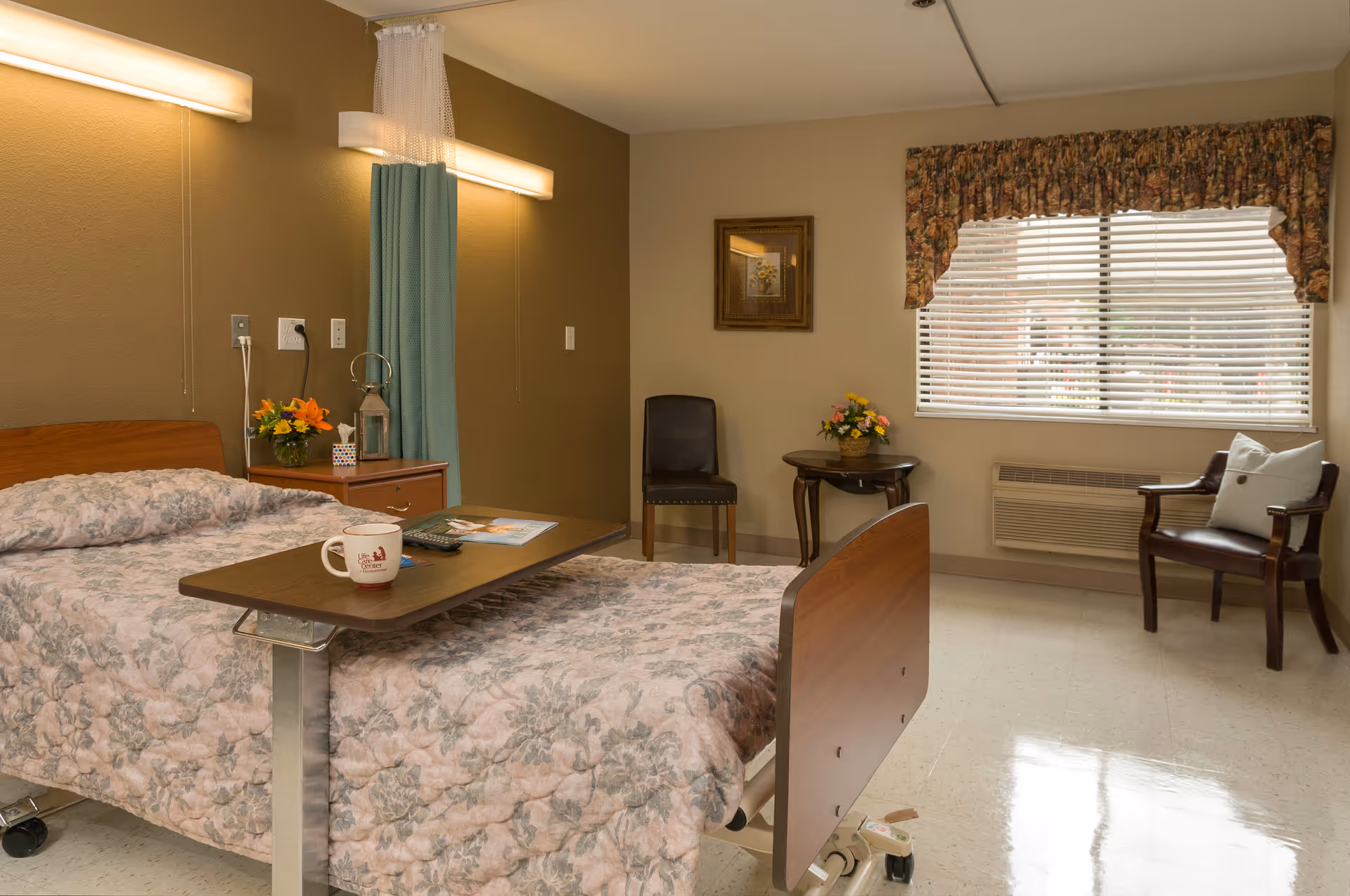 A senior living facility bedroom with a hospital-style bed covered in a floral quilt. A wooden overbed table holds a mug, remote control, and magazine. There is a nightstand with a flower arrangement and a lantern, a chair, a small round table with another flower arrangement, and a window with blinds and floral valance. The room has beige walls and tiled flooring.