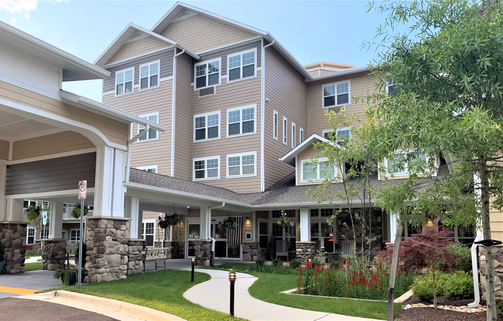 Front entrance of a multi-story senior living building with a covered drop-off, stone columns, landscaped walkway and hanging plants.