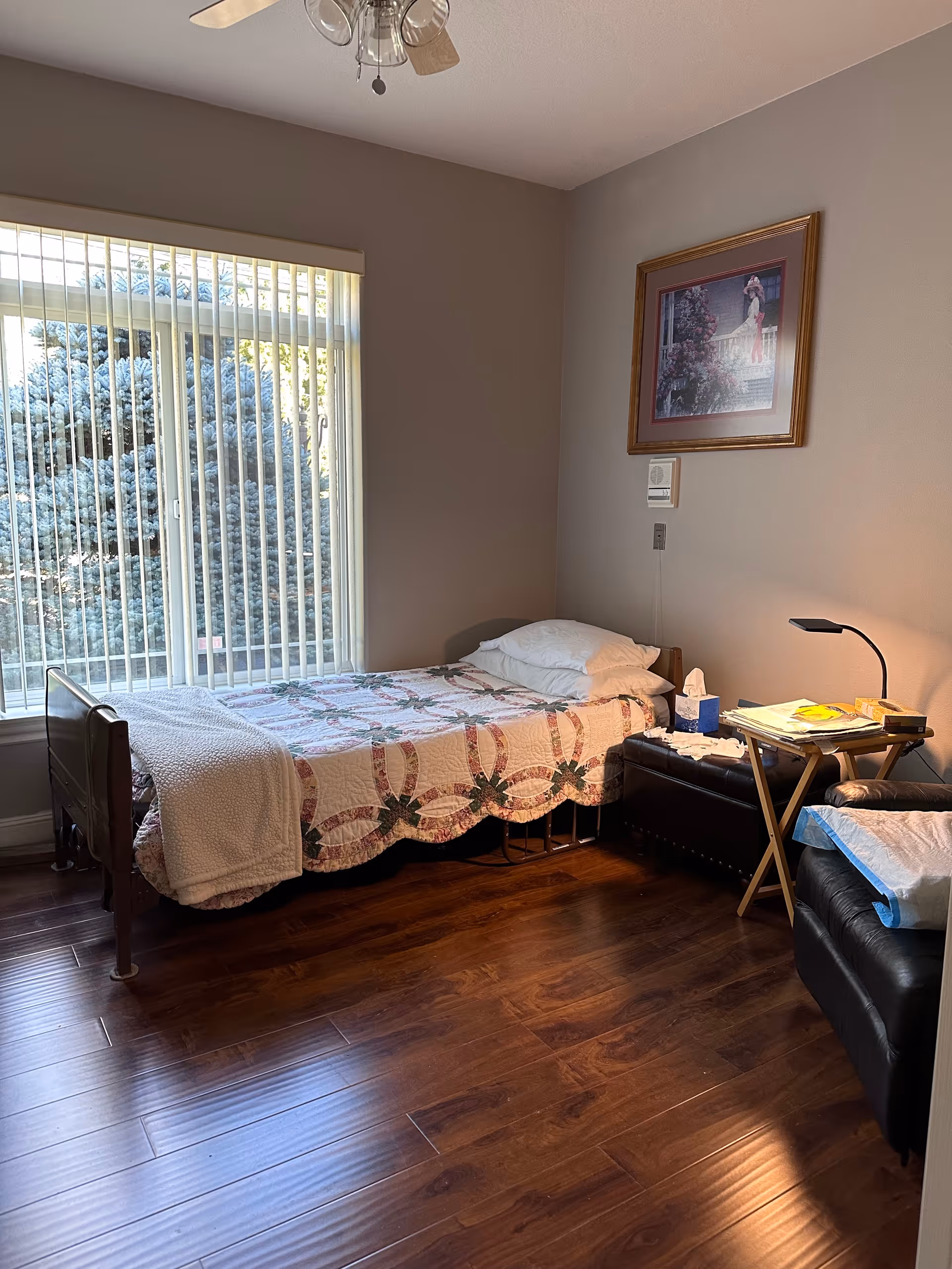 A single bed with a patterned quilt and white pillows is placed next to a large window with vertical blinds. Beside the bed is a small table with a lamp, tissues, and some books or magazines. A black leather chair with a white cloth draped over the arm is also visible. The room has wooden flooring and a framed picture hanging on the wall above the bed.