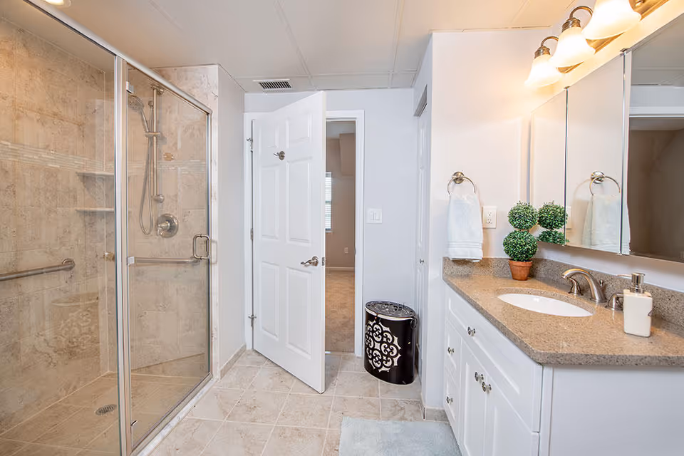 A clean and modern bathroom featuring a glass-enclosed shower with beige tile walls, a white vanity with a granite countertop, a sink, a soap dispenser, a small potted plant, a towel hanging on a ring, and a partially open white door leading to another room.