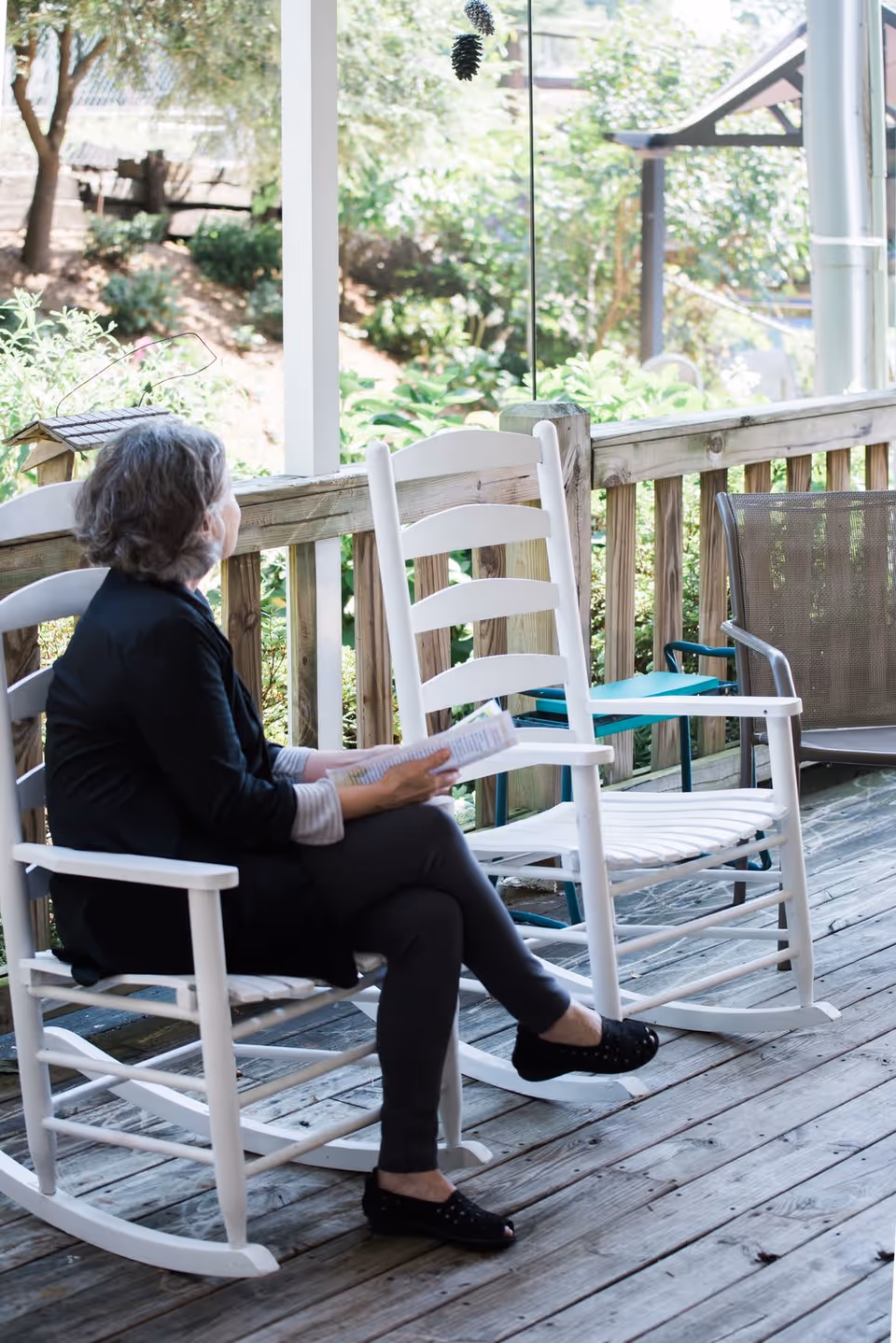 An elderly woman sitting on a white wooden rocking chair on a wooden porch, holding a book and looking out towards a garden with greenery and trees.