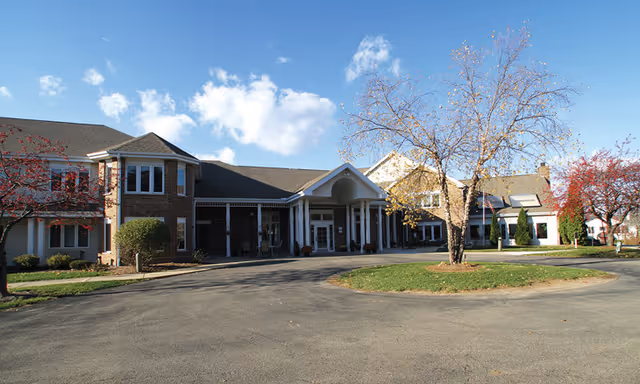 Front exterior view of a senior living facility named Eden Vista Madison, featuring a large building with a covered entrance, brick and siding exterior, surrounded by trees with autumn foliage and a clear blue sky.