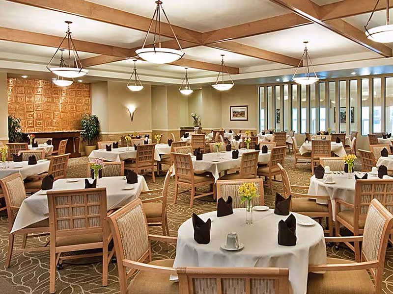 A spacious dining room with multiple round and rectangular tables covered with white tablecloths, each set with black folded napkins, cups, and small vases of yellow flowers. The room features wooden chairs, a patterned carpet, warm lighting from ceiling fixtures, and a decorative wall panel with plants in the background.