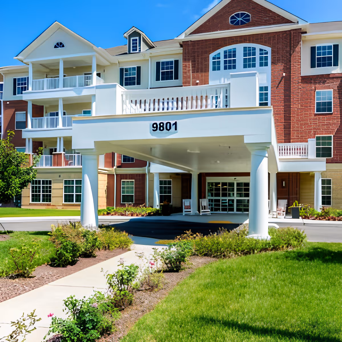 Front exterior view of a multi-story senior living facility with a covered entrance displaying the number 9801. The building features a combination of brick and light-colored siding, multiple windows, balconies, and a well-maintained lawn with shrubs and flowers in the foreground under a clear blue sky.