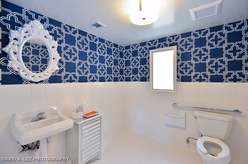 A clean and modern bathroom with white fixtures including a sink, toilet, and a small cabinet. The walls are decorated with white tiles on the lower half and a blue patterned wallpaper on the upper half. A decorative white-framed mirror hangs above the sink, and there is a window letting in natural light. A grab bar is installed next to the toilet for accessibility.