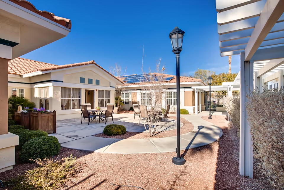 Sunny courtyard with patio tables and chairs, lamp posts, landscaping, and single-story buildings with tiled roofs.