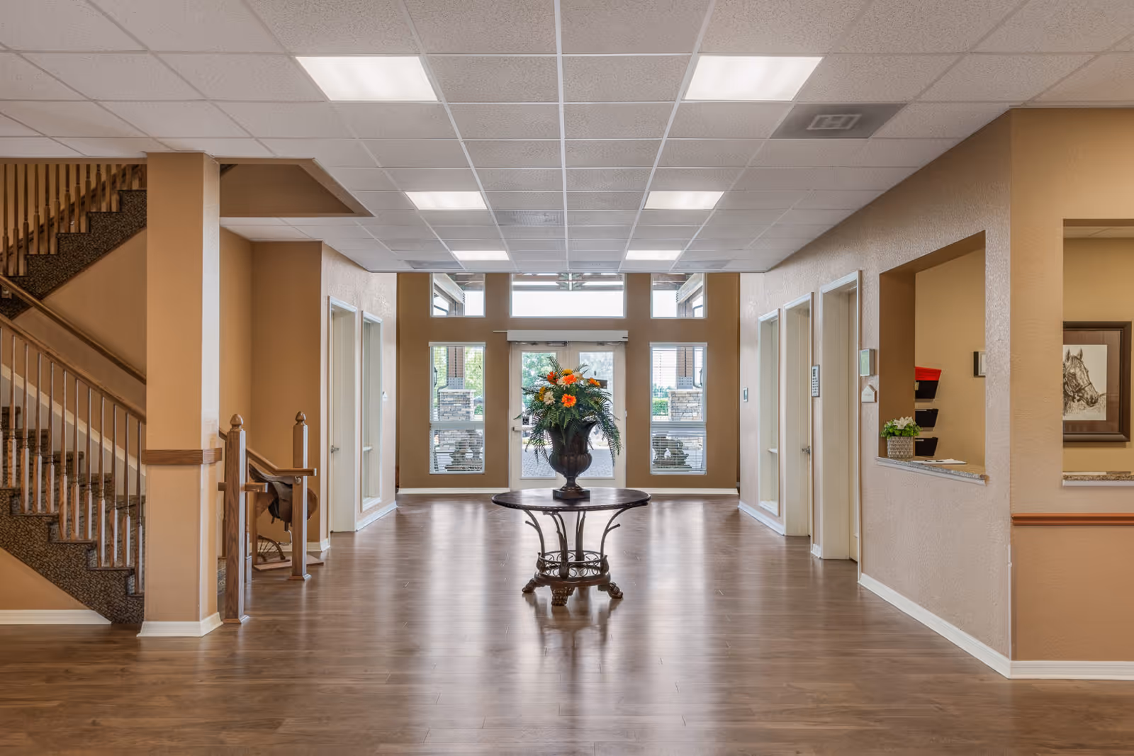 A spacious and well-lit interior hallway with wood flooring and beige walls. In the center, there is a round wooden table with a large floral arrangement featuring orange and white flowers. To the left, there is a staircase with wooden railings. On the right side, there is a reception window with a small plant and some wall-mounted organizers. At the far end, glass doors and windows allow natural light to enter, showing a glimpse of the outdoor area.