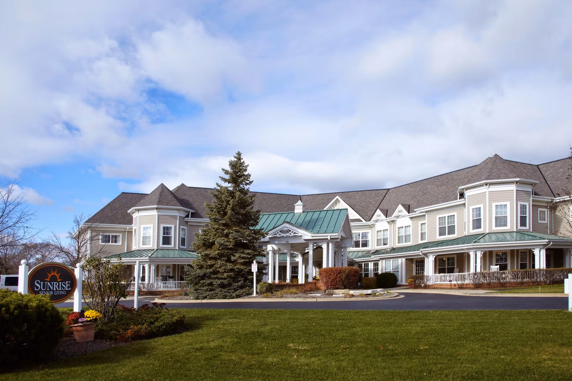Exterior view of Sunrise of Buffalo Grove senior living facility, showing a large two-story building with gray siding, white trim, and green metal roofing accents. The building features multiple windows, a covered entrance with white columns, and a well-maintained lawn with shrubs and a tall evergreen tree. A sign near the entrance reads 'Sunrise Senior Living.'