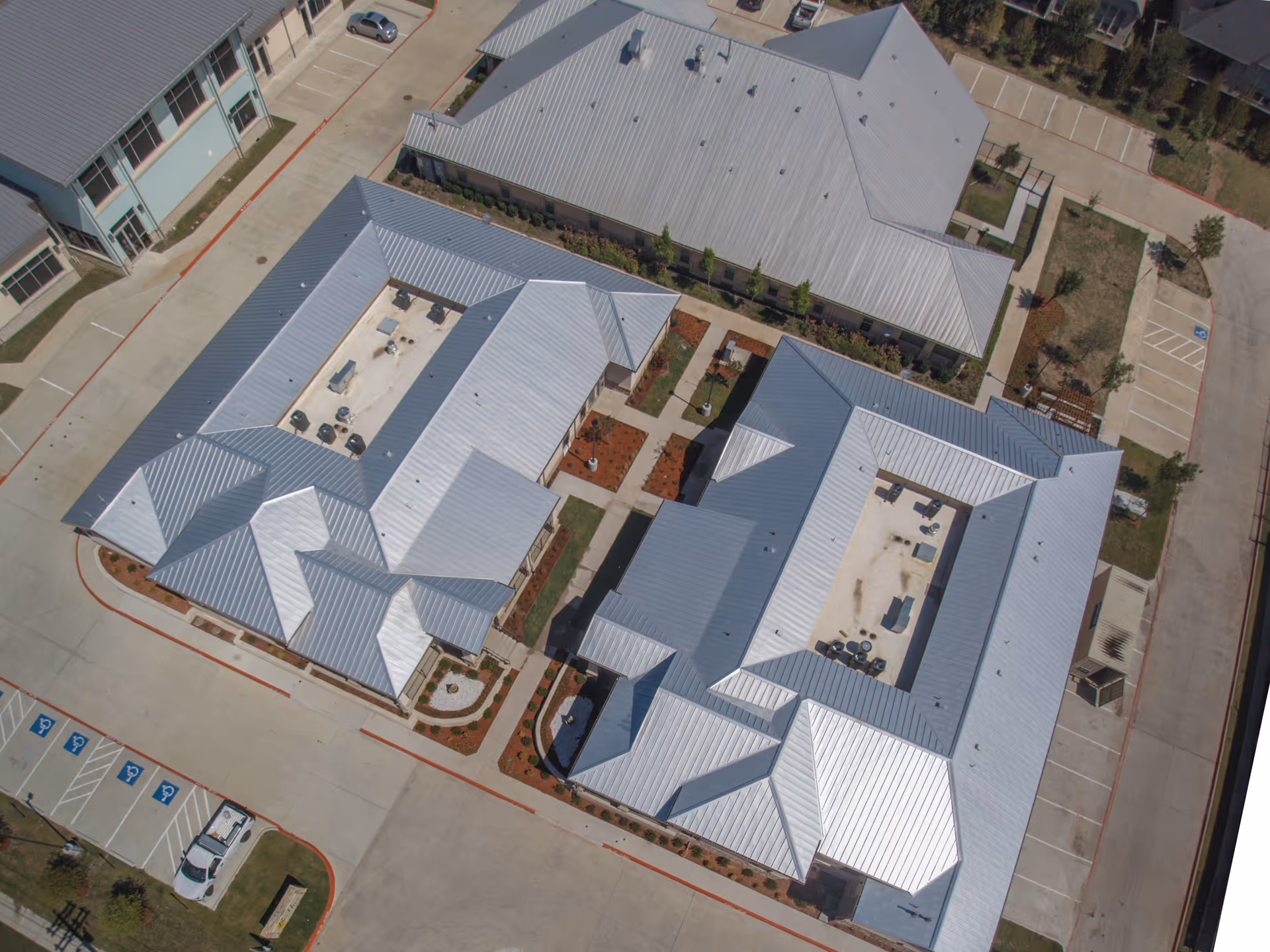 Aerial view of BeeHive Homes of Frisco showing multiple connected buildings with metal roofs, surrounding parking lots with marked handicap spaces, and landscaped areas with pathways.