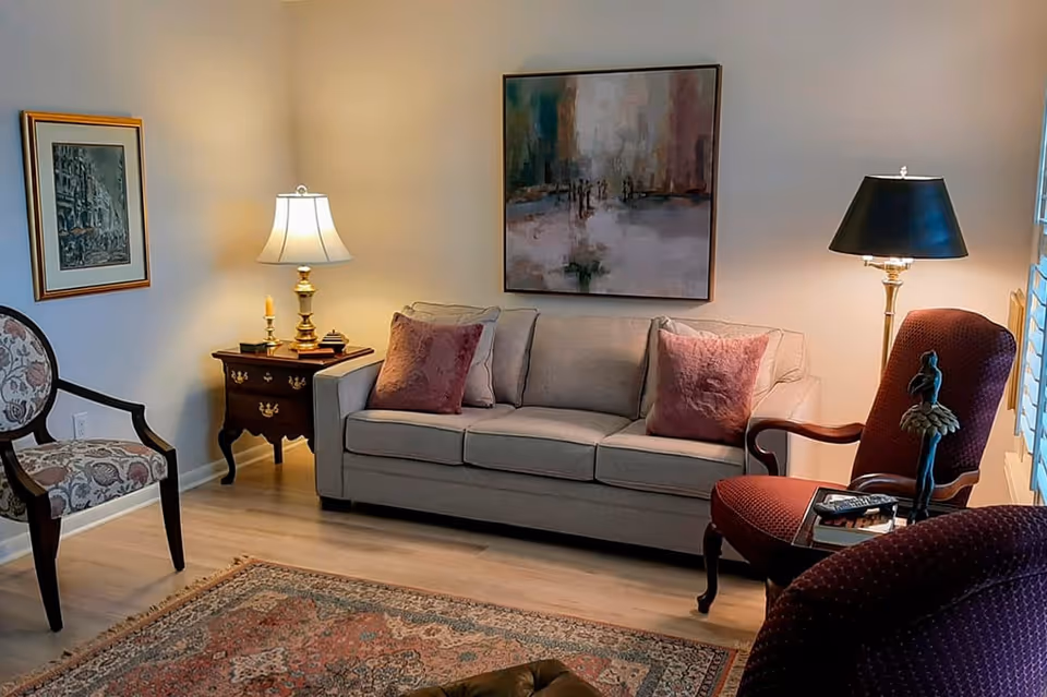 A cozy living room with a beige sofa adorned with two mauve pillows, a wooden side table with a lamp and candles, two upholstered armchairs, a floor lamp, a framed painting on the wall, and a patterned area rug on a wooden floor.
