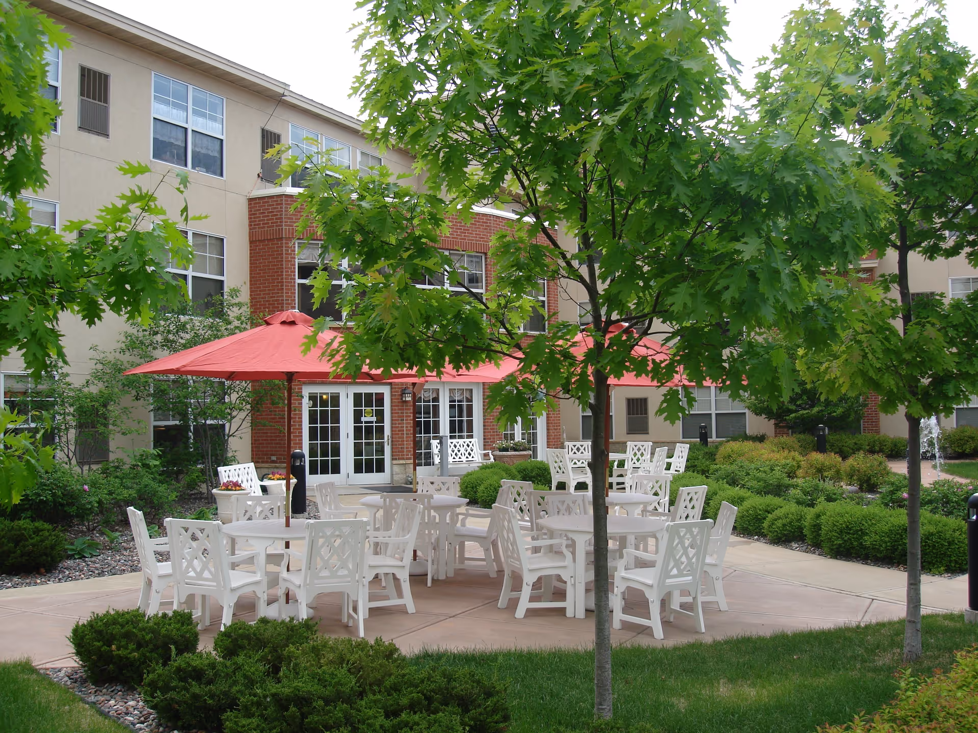 Outdoor patio area at The Pines Senior and Assisted Living with white tables and chairs arranged under red umbrellas, surrounded by green trees and bushes, with the building visible in the background.