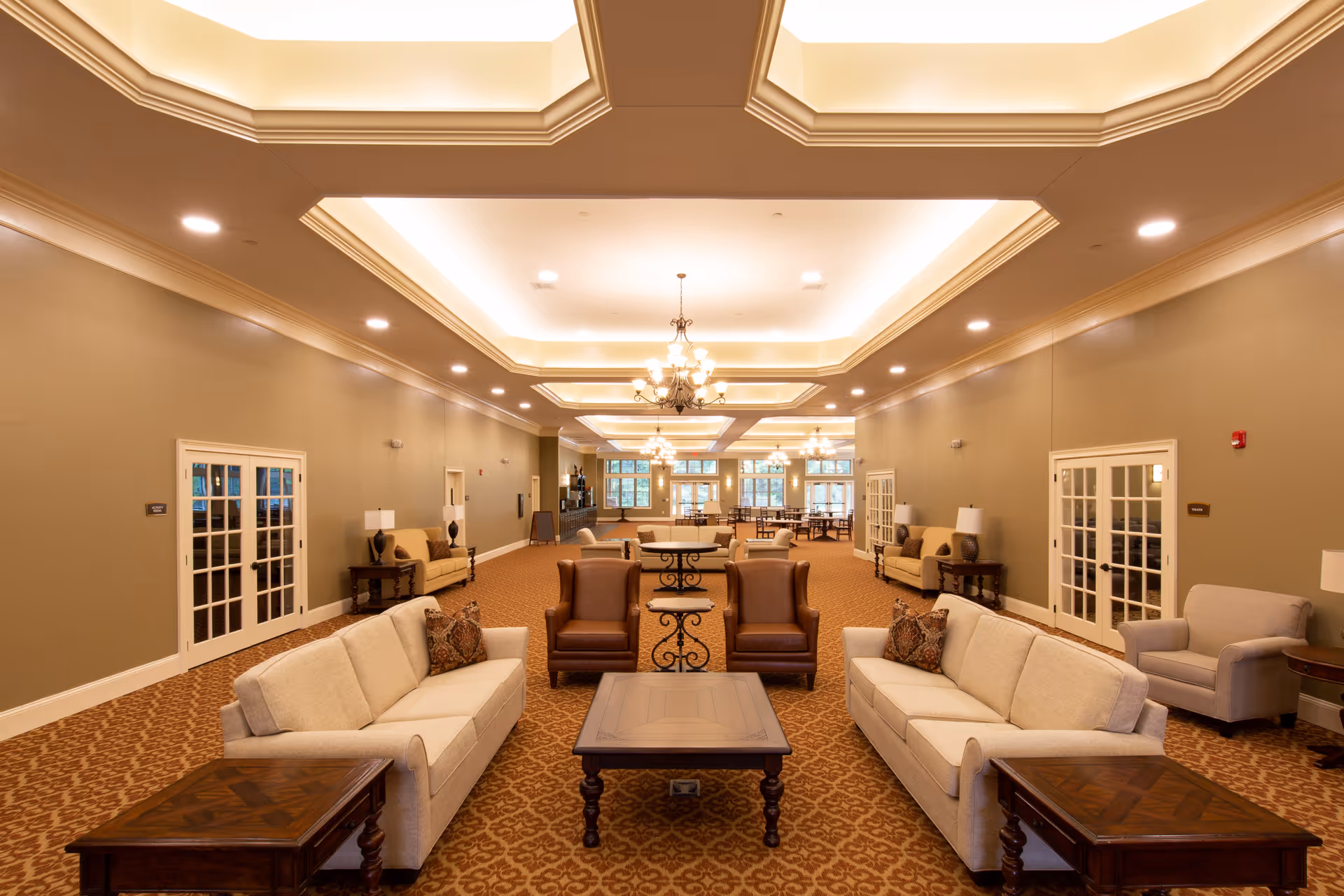 A spacious and well-lit common area in an assisted living facility featuring beige sofas, brown armchairs, wooden coffee tables, and side tables with lamps. The room has a patterned carpet, neutral-colored walls, and decorative ceiling lighting with chandeliers. French doors line the walls, and large windows are visible in the background.