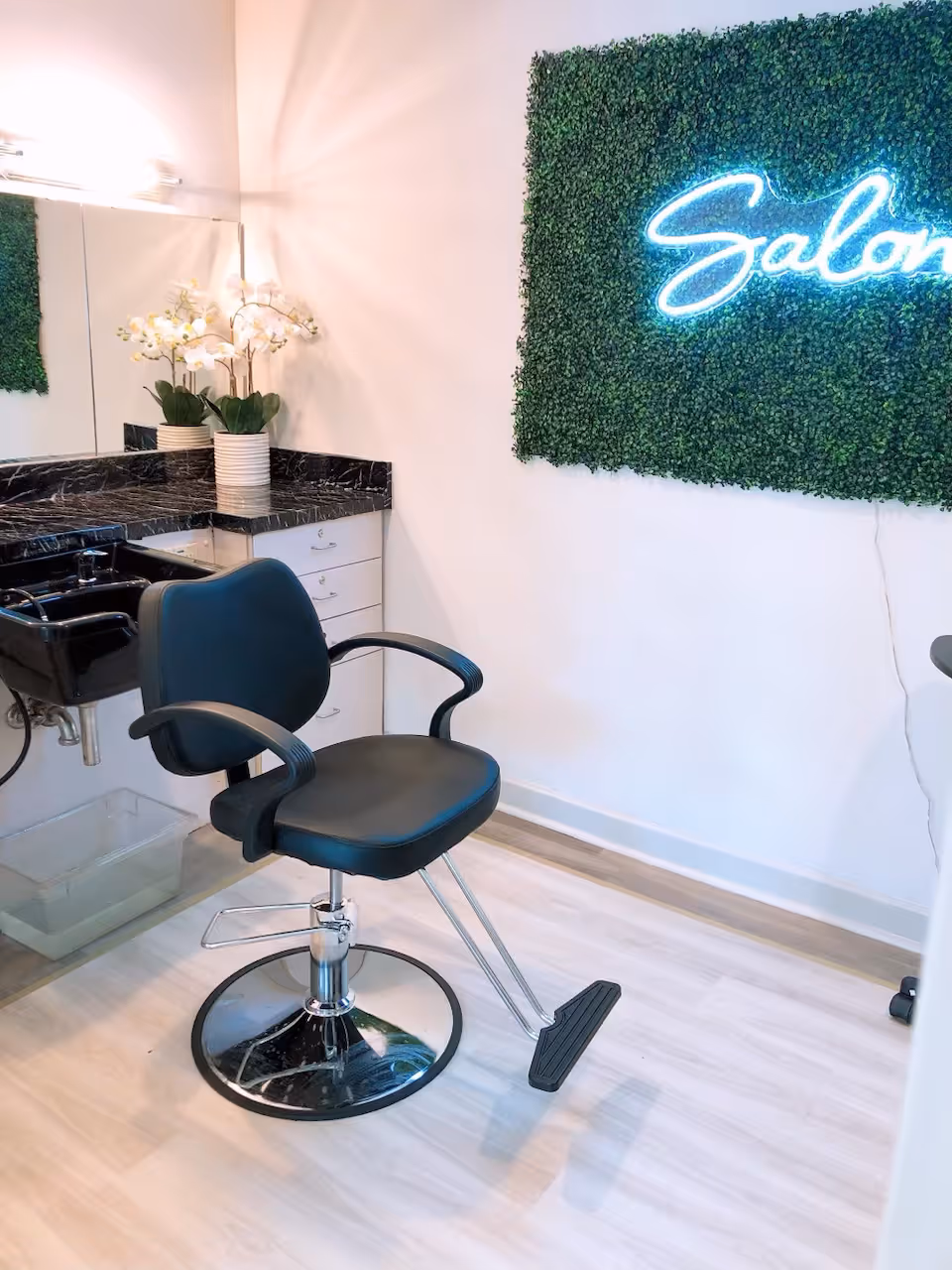 A salon chair in front of a black marble countertop with a sink, white cabinets, and a mirror. There is a decorative green wall panel with a neon sign that says 'Salon'. A potted orchid plant is placed on the countertop.