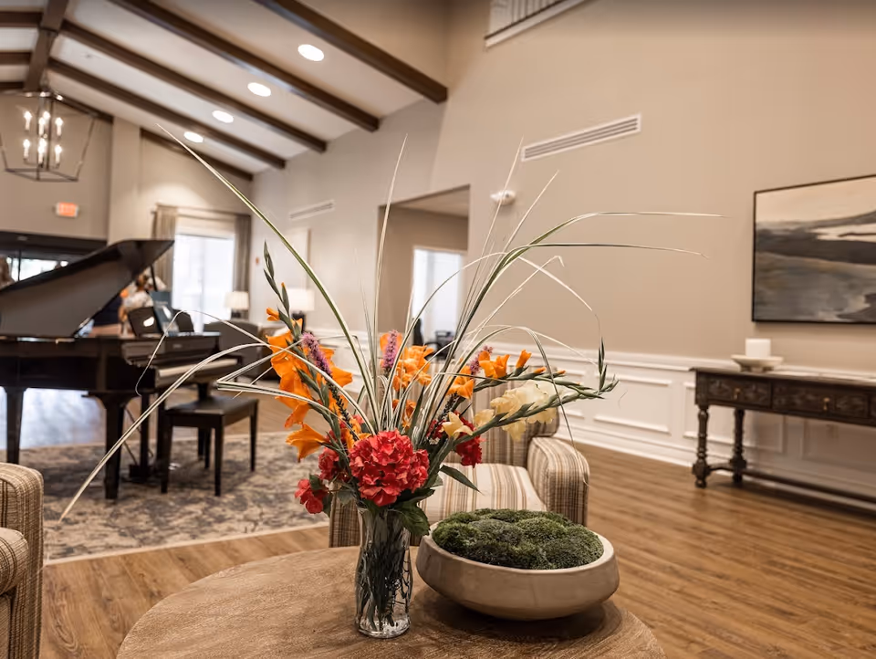 Well-lit communal lounge with a floral arrangement on a wooden table, a grand piano, seating, and a decorative console table.