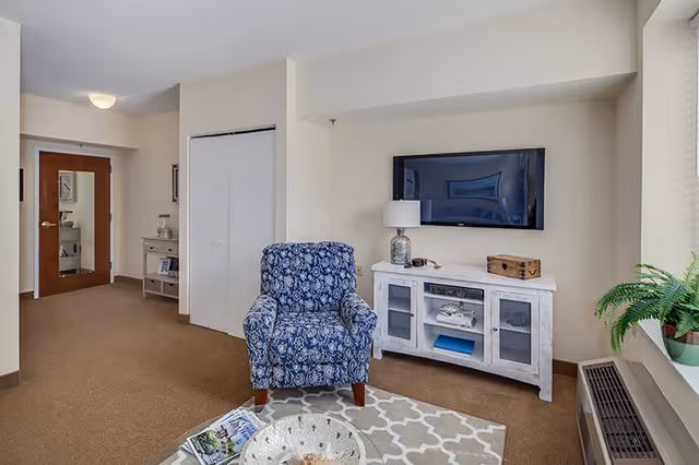 A cozy living room area with a blue patterned armchair placed on a gray and white patterned rug. A white wooden cabinet with glass doors holds decorative items and a lamp, with a flat-screen TV mounted on the wall above it. There is a window with blinds and a green potted plant on the windowsill. The room has beige walls and carpet, with a wooden door and a small console table visible in the background.
