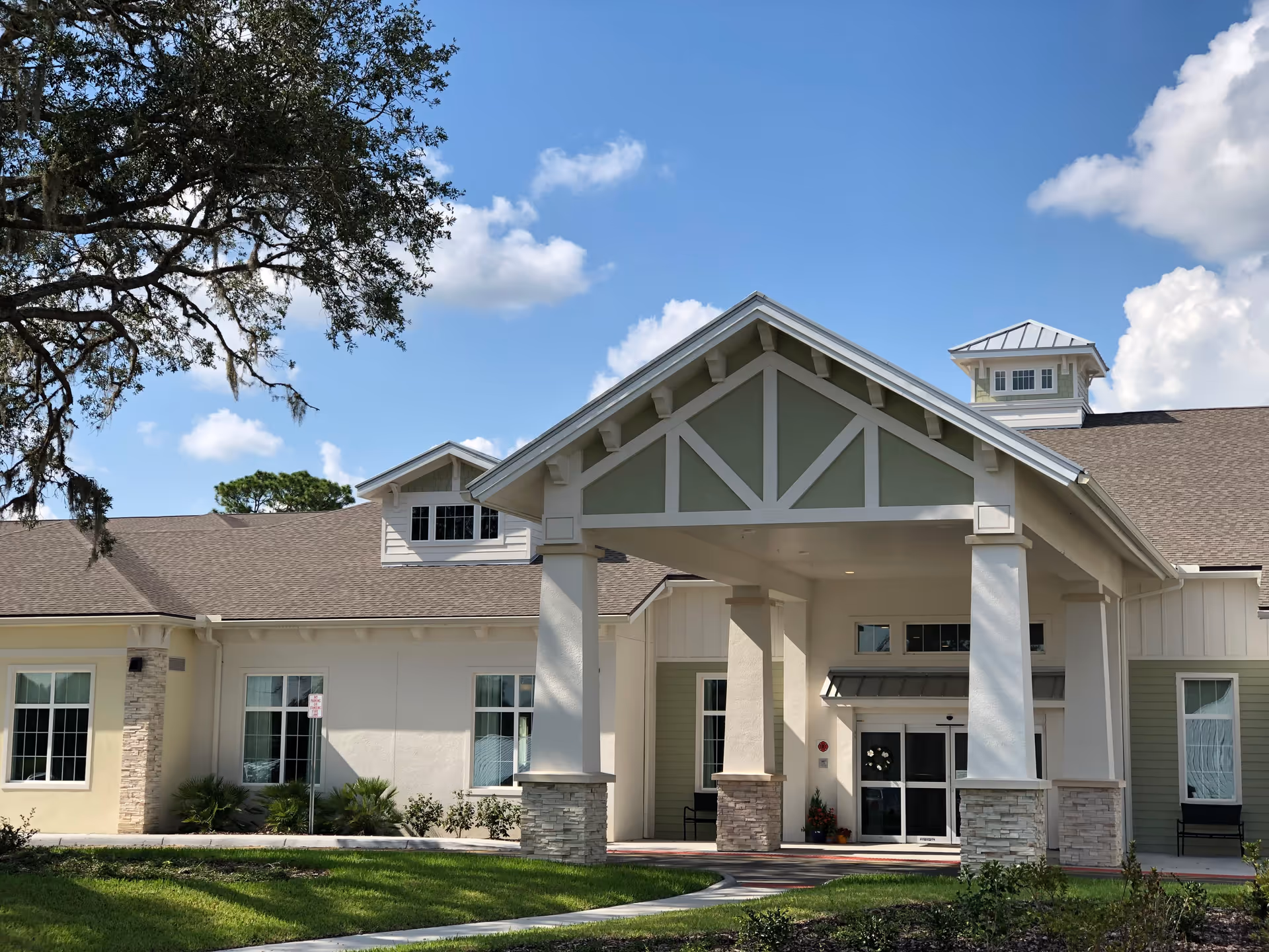 Exterior view of a senior living facility building with a covered entrance supported by columns, beige and light green walls, and a brown shingled roof under a blue sky with some clouds. There are windows and some landscaping with grass and small plants in front.