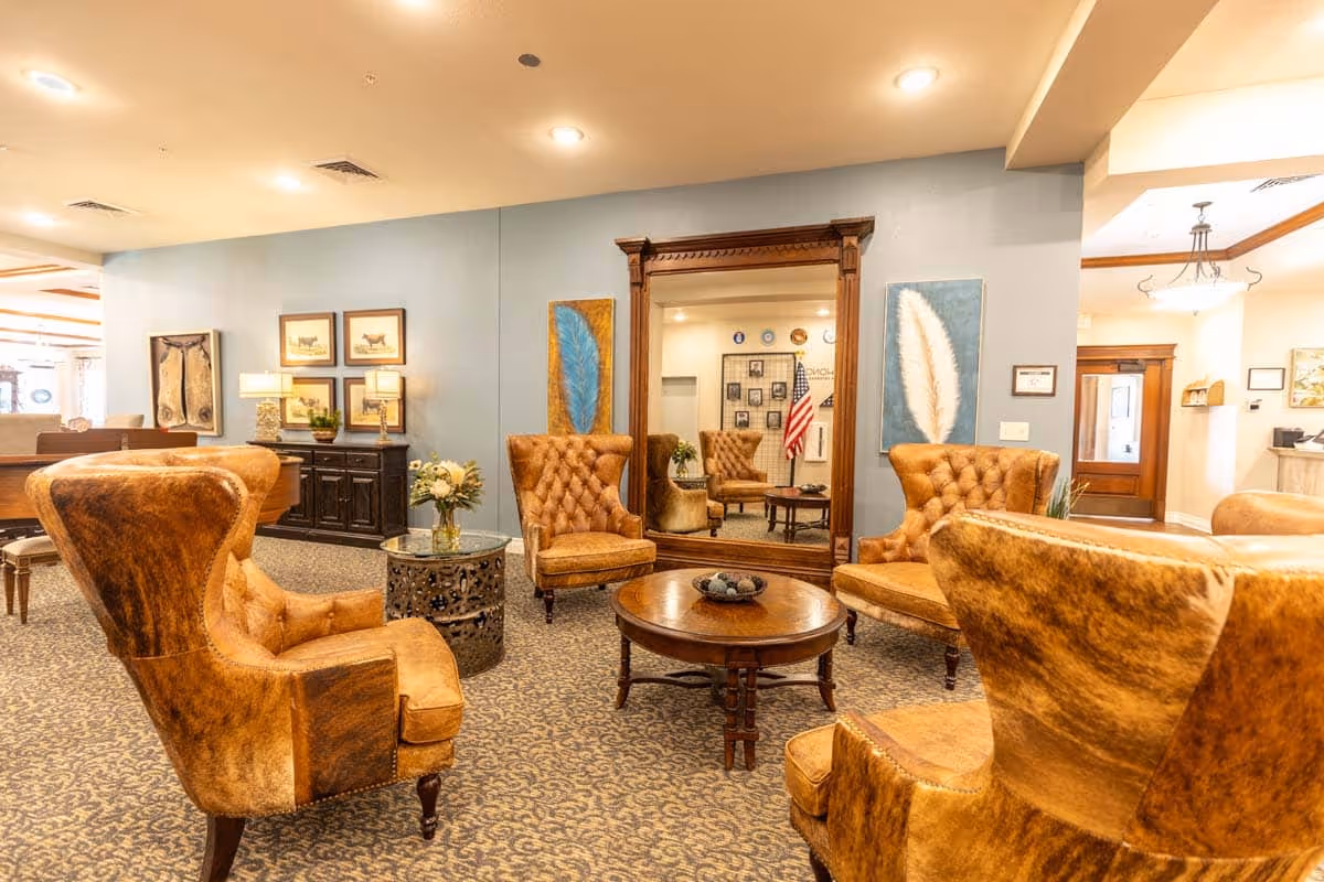 A cozy seating area in a senior living facility with four brown leather armchairs arranged around a round wooden coffee table. The room features a large ornate mirror on the wall, decorative paintings, a side table with a flower vase, and warm lighting. The carpet has a patterned design, and the overall ambiance is inviting and comfortable.