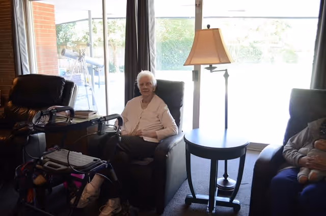 An older woman seated in a recliner in a living room area beside a walker, small round table, and floor lamp in front of sliding glass doors.
