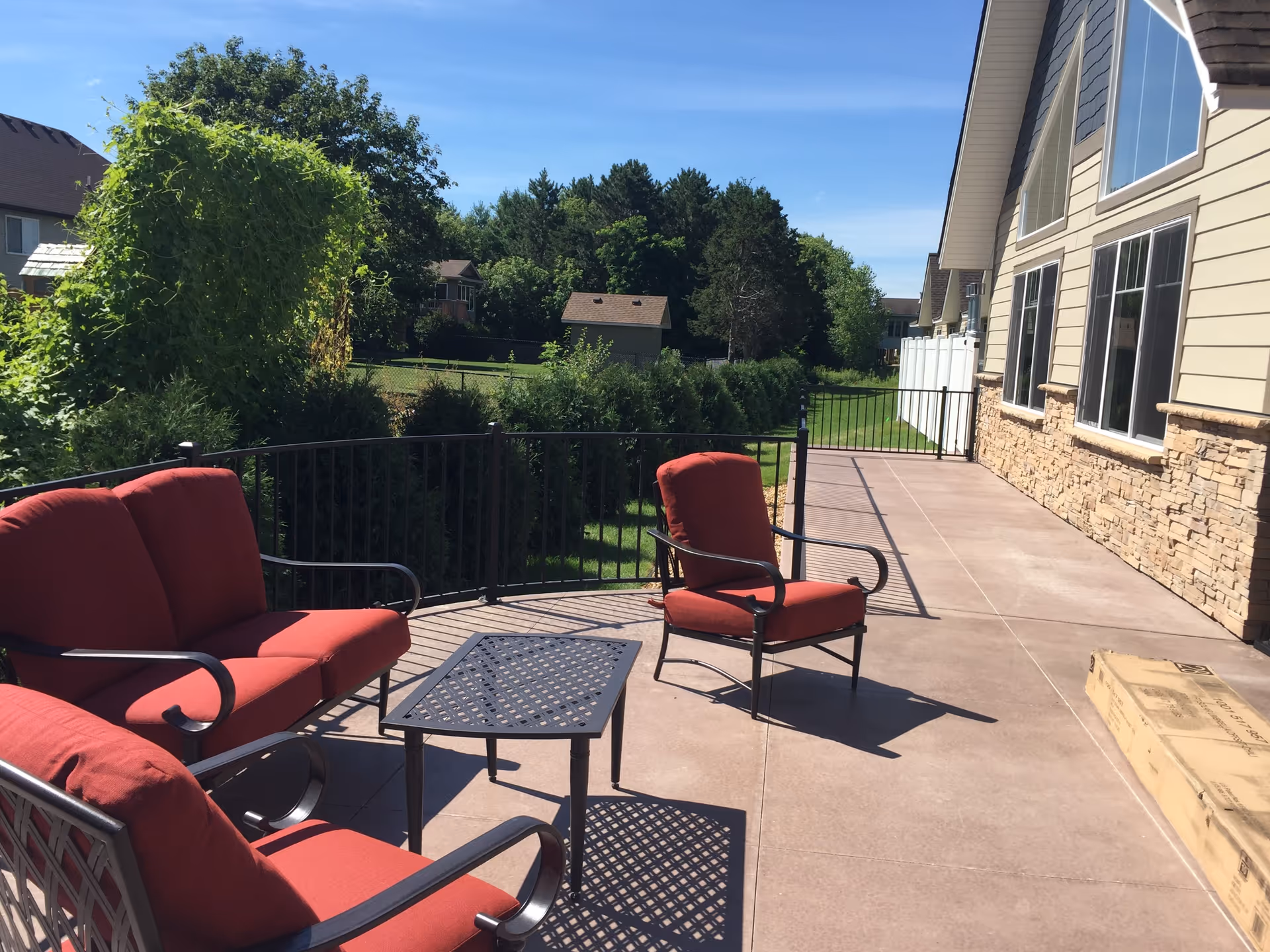 Outdoor patio with red-cushioned chairs and a metal table beside a building overlooking a grassy yard and trees.