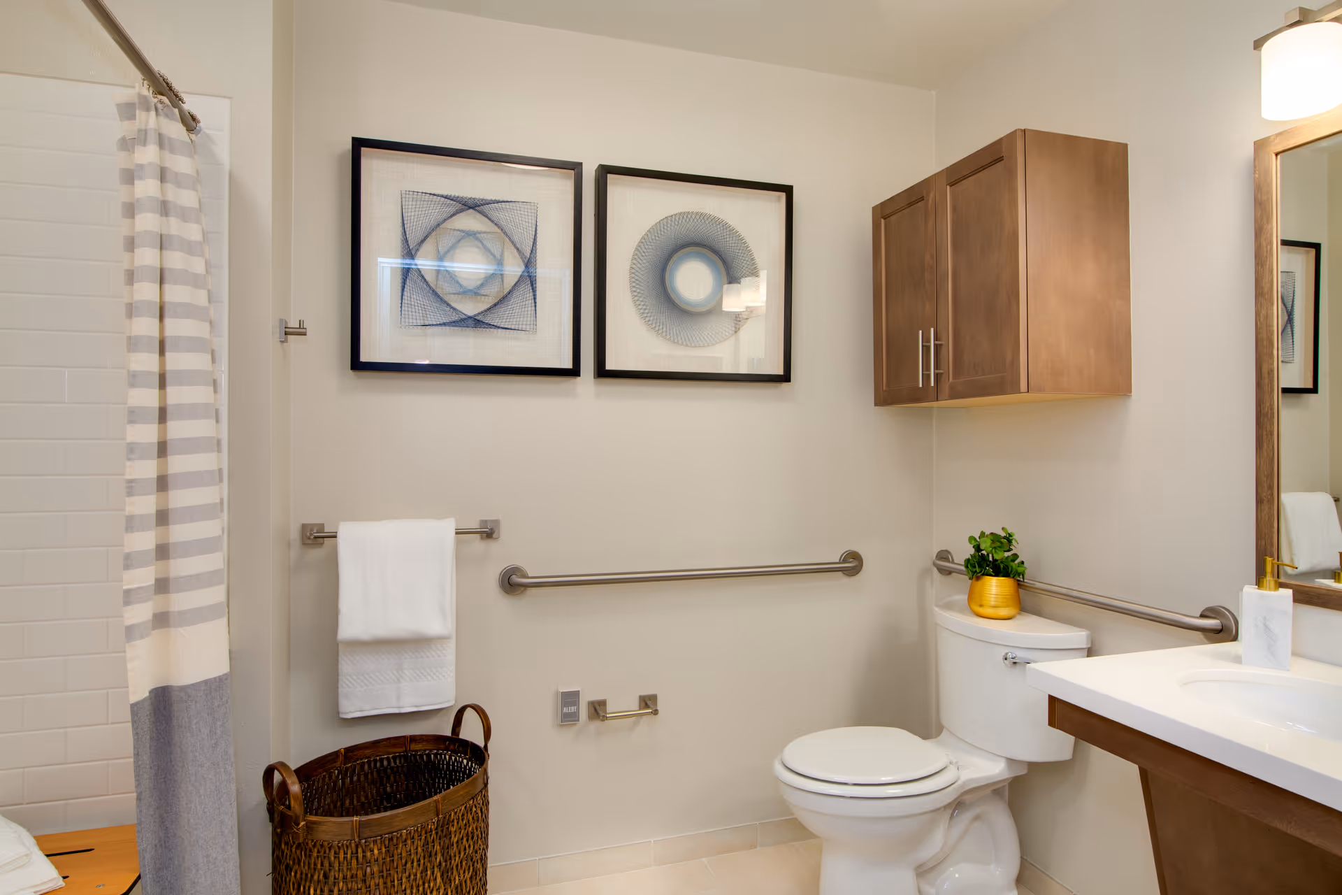 A clean and modern bathroom featuring a white toilet with a small green plant in a yellow pot on the tank, a wooden cabinet mounted on the wall above the toilet, a white countertop with a sink and soap dispenser, a large mirror, and two framed abstract art pieces on the wall. There is a towel rack with a white towel, a striped shower curtain, and a woven basket on the floor.