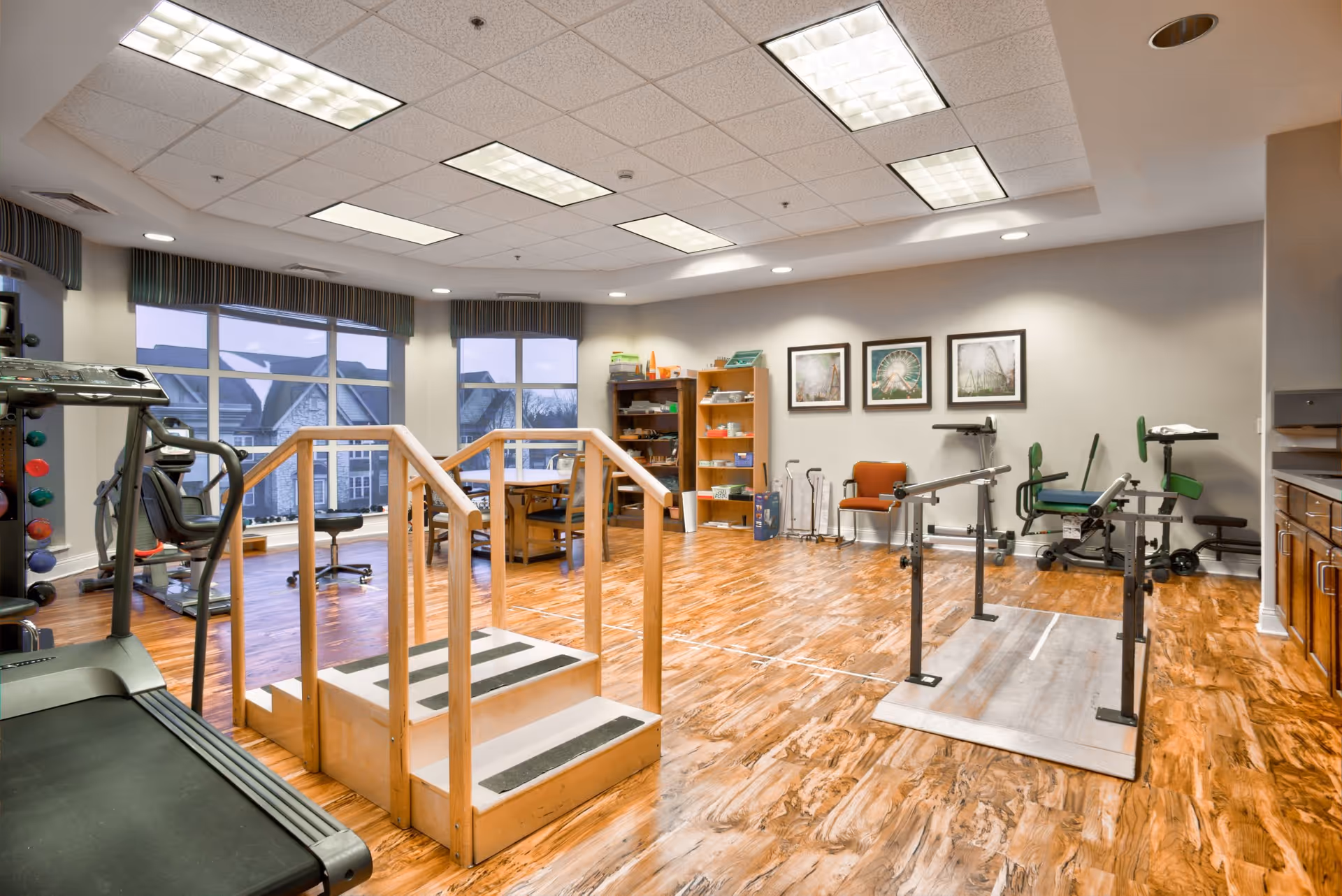 A spacious physical therapy or rehabilitation room with wooden flooring, large windows, and exercise equipment including a treadmill, hand weights, parallel bars, and therapy chairs. There are framed pictures on the wall and shelves with various supplies.