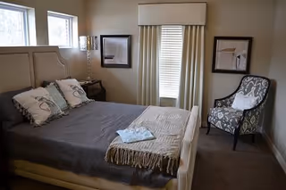 A bedroom with a large bed covered in a gray blanket and decorative pillows. There is a folded towel and a beige throw blanket on the bed. The room has three small windows on one wall, a window with closed blinds and curtains on another wall, two framed pictures, a patterned armchair with a white pillow, and a small bedside table with a lamp.