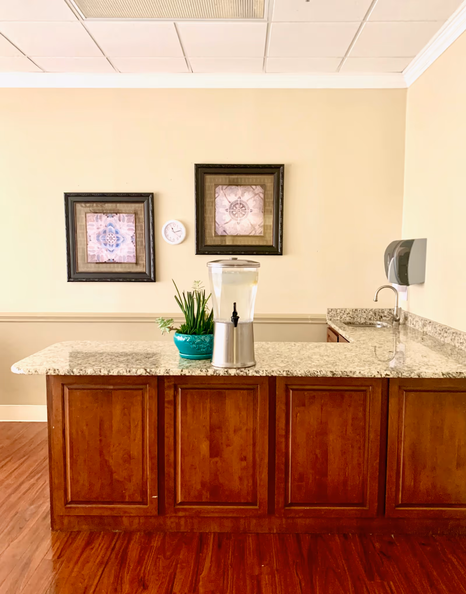 Interior view of a room with a granite countertop island featuring a water dispenser and a green potted plant. The countertop has wooden cabinets underneath. On the wall behind the counter, there are two framed decorative pictures and a small round clock. A wall-mounted soap dispenser and a small sink are visible to the right.