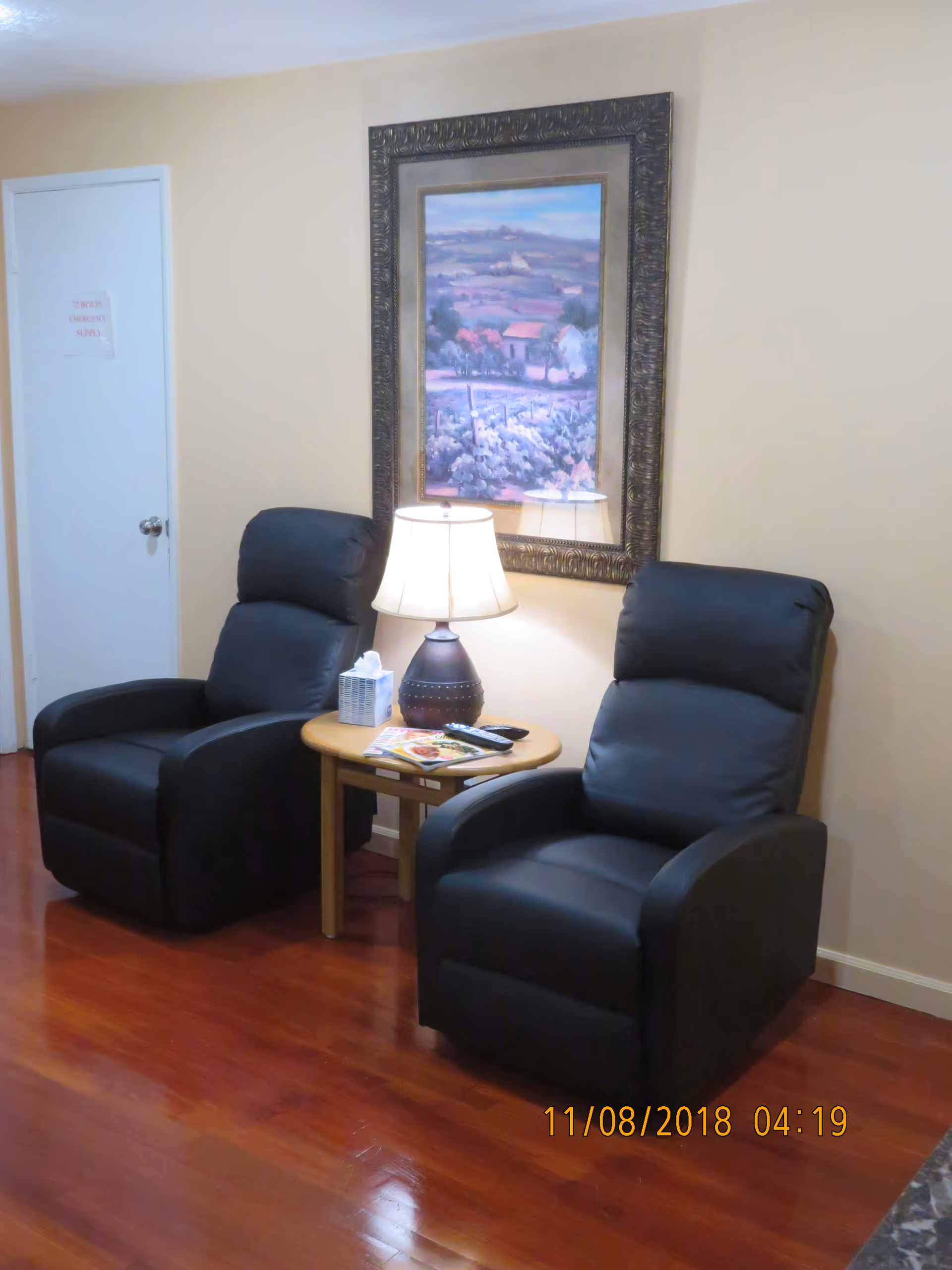 Two black recliner chairs flanking a small round table with a lamp beneath a framed landscape painting on a hardwood floor.