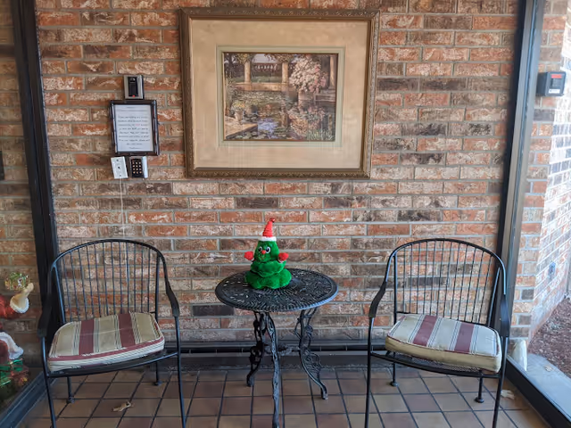A small outdoor seating area with two metal chairs with striped cushions and a round metal table between them. On the table is a green plush toy wearing a red and white Santa hat. The background is a brick wall with a framed painting of a garden scene hanging above the table. There is a keypad and a small sign mounted on the wall to the left.