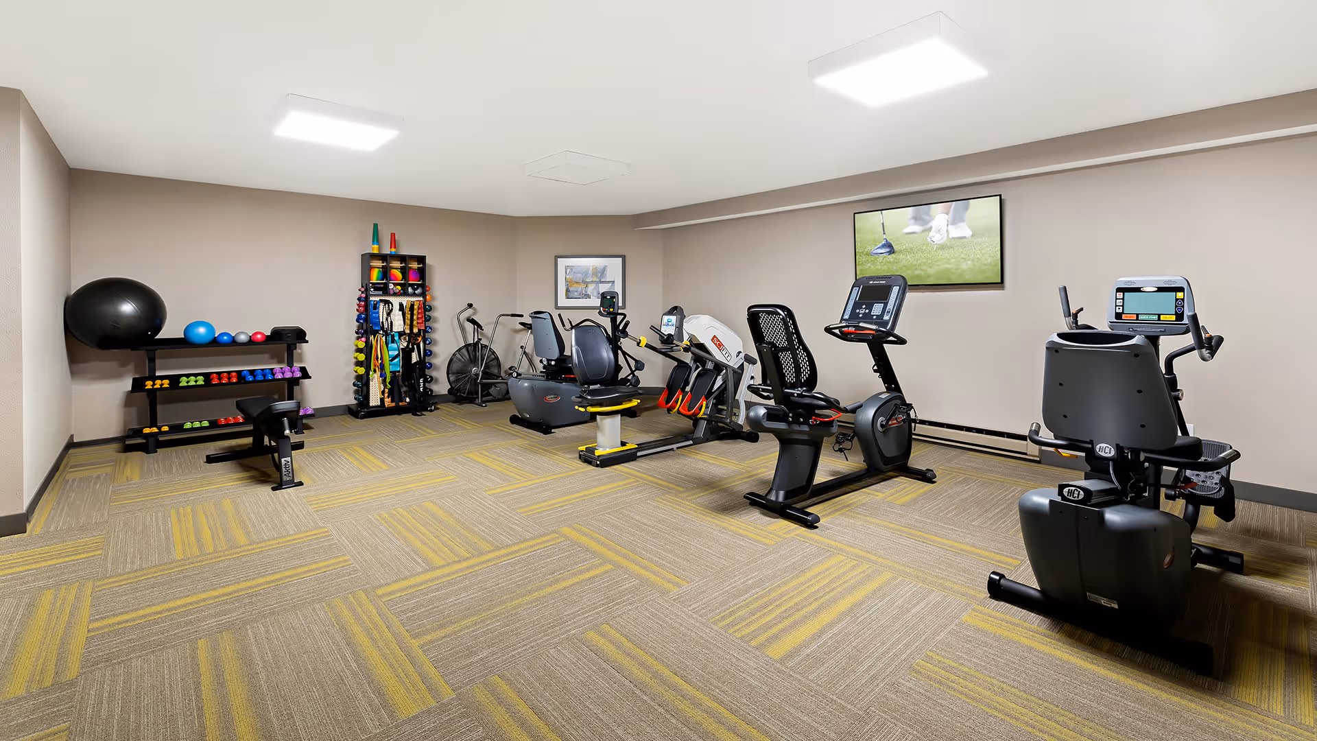 A fitness room with exercise equipment including stationary bikes, a rowing machine, and various weights and exercise balls neatly arranged on racks. A television is mounted on the wall showing a golf scene. The room has beige walls and patterned carpet flooring in shades of yellow and gray.