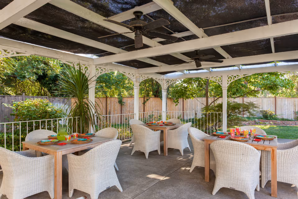 Outdoor covered patio area with white wicker chairs and wooden tables set with colorful plates and glasses, surrounded by greenery and a wooden fence.