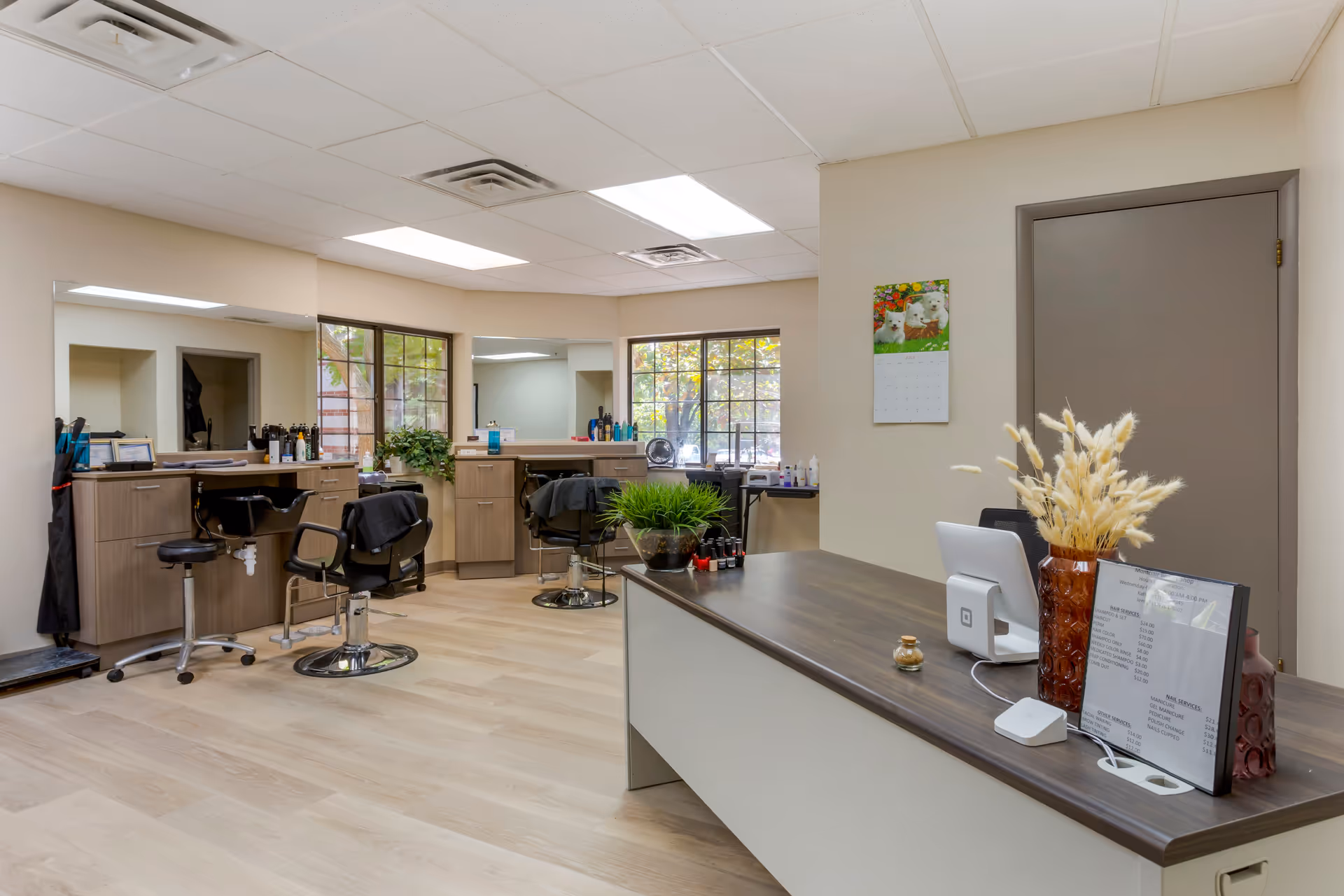Interior view of a salon area with styling chairs, mirrors, and hair care products. There is a reception desk in the foreground with a vase of dried flowers, a payment terminal, and a price list. The room has large windows letting in natural light and light-colored wood flooring.