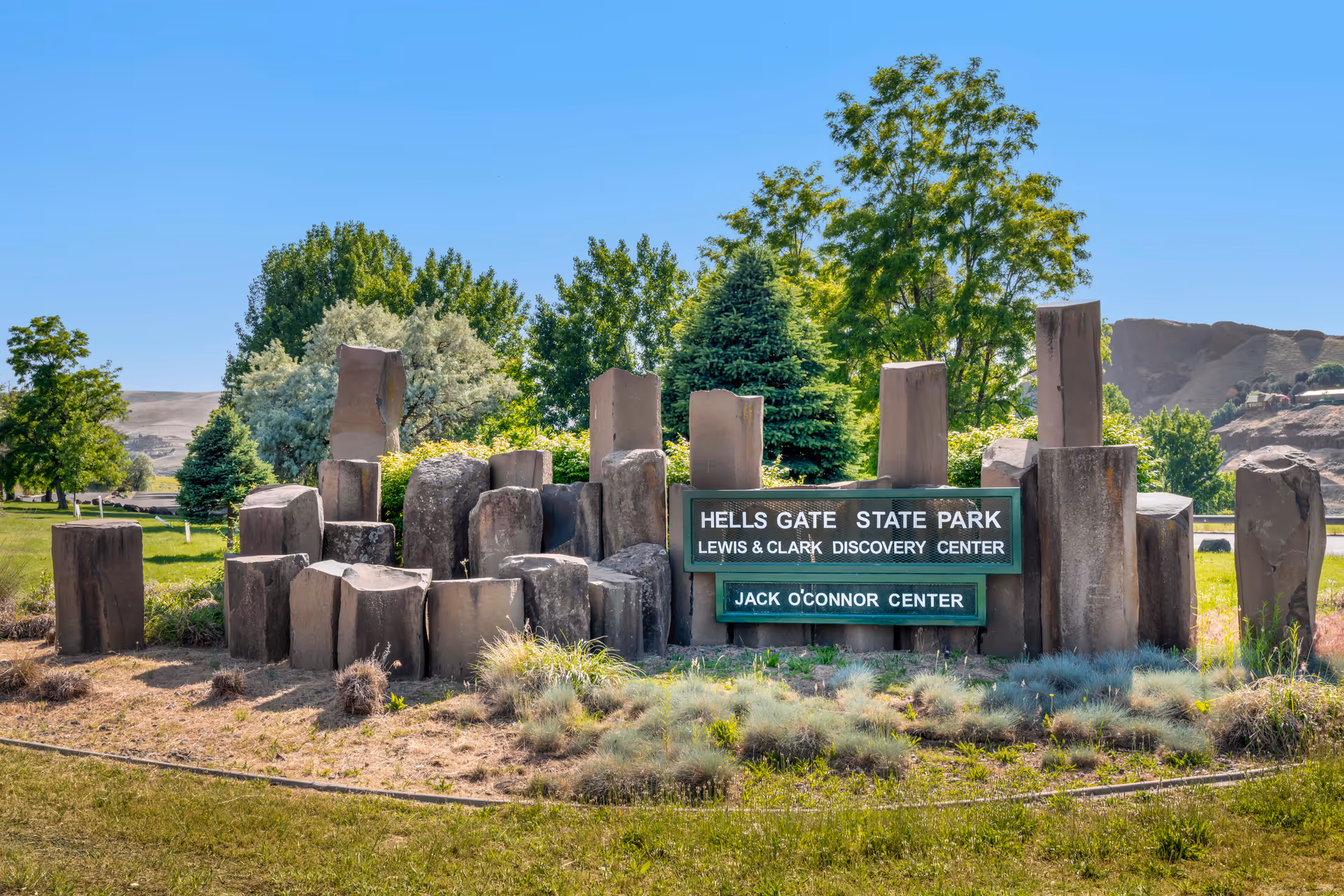 Stone monument sign for Hells Gate State Park, Lewis & Clark Discovery Center, and Jack O'Connor Center surrounded by grass, shrubs, and trees under a clear blue sky.