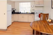 Small kitchenette with light wood cabinets, white appliances, and a wooden dining table in the foreground.