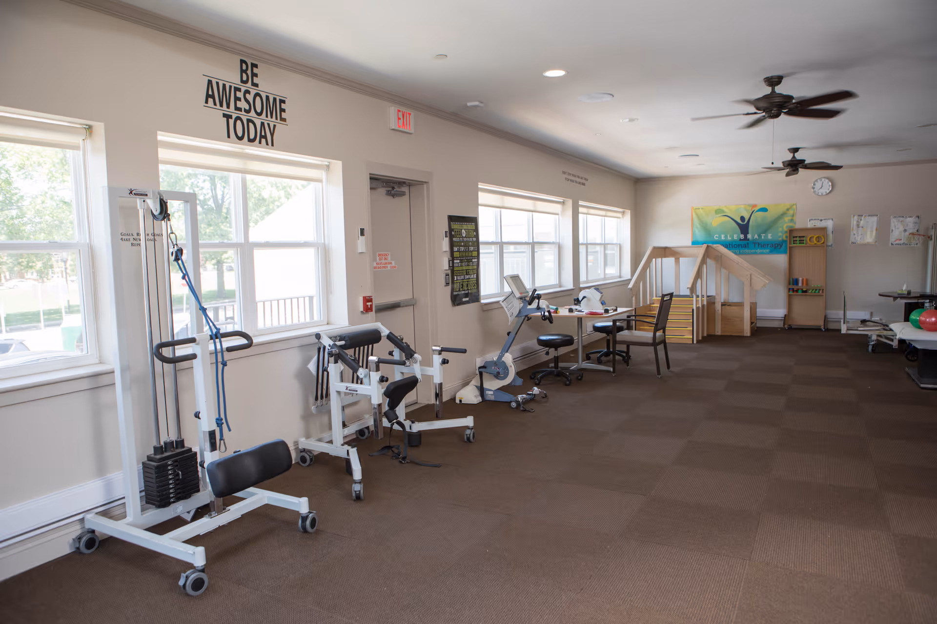A spacious therapy room with exercise equipment including a stationary bike, weight machines, and therapy stairs. The room has large windows letting in natural light, ceiling fans, and motivational text on the wall that reads 'BE AWESOME TODAY'.