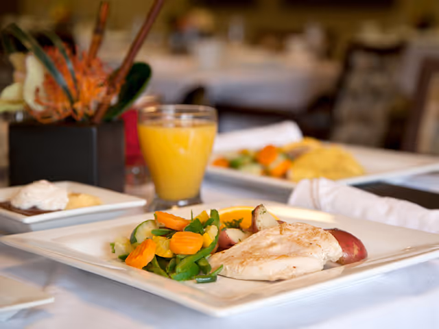 A plated meal consisting of grilled chicken breast, mixed steamed vegetables including carrots and green beans, and roasted red potatoes on a white square plate. A glass of orange juice and a small dessert plate with a dollop of whipped cream are also visible on the table. The setting appears to be a dining area with white tablecloths and blurred background tables and chairs.